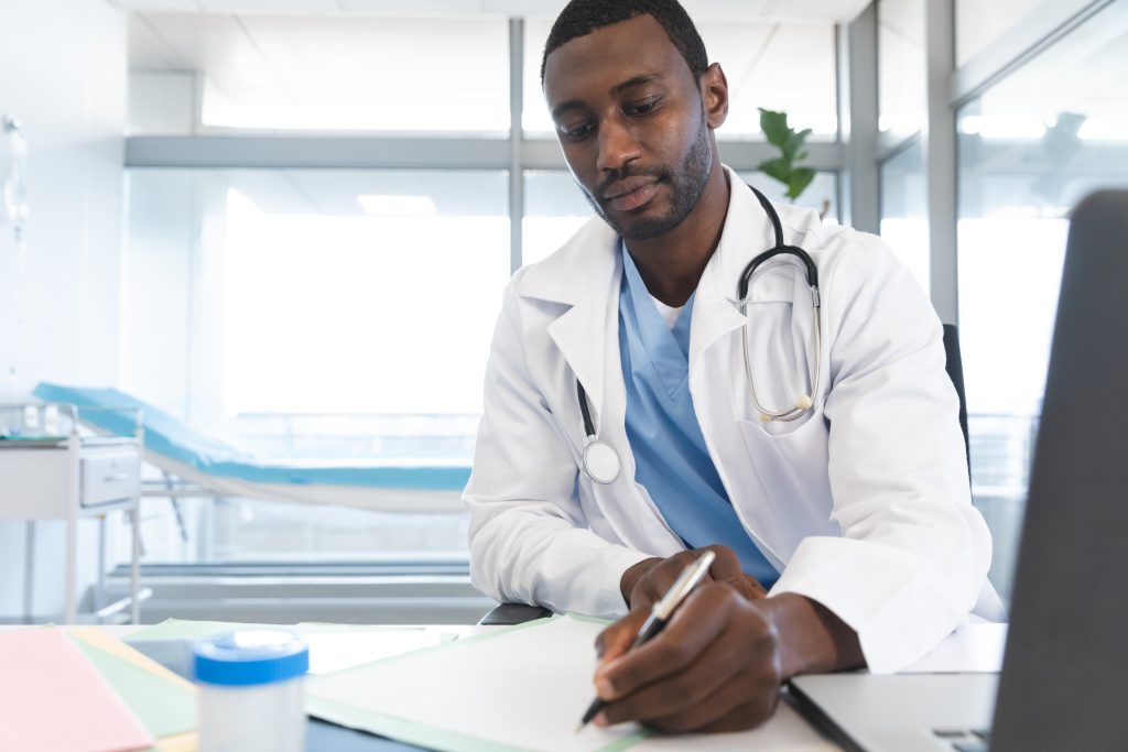 Male doctor wearing lab coat and stethoscope, sitting at desk, taking notes. Hospital, medicine, healthcare and work, unaltered.